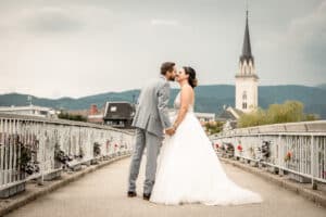 Braut und Bräutigam küssen sich auf einer malerischen, mit Blumen geschmückten Brücke. Die Braut trägt ein weißes Kleid und der Bräutigam einen grauen Anzug. Im Hintergrund dieser perfekten Hochzeit runden malerische Berge, Gebäude und eine Kirche mit einem hohen Kirchturm ihren magischen Tag ab.