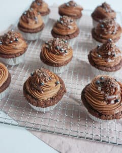 Zwölf Schokoladen-Cupcakes mit Schokoladenglasur und kleinen silbernen Kugeln und Schokoladenraspeln garniert, sind elegant auf einem Kuchengitter mit einem hellrosa Tuch darunter arrangiert – perfekt, um Ihrer Hochzeit eine süße Note zu verleihen.