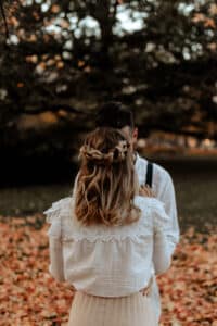 Inmitten von herbstlichen Blättern steht ein Paar eng beieinander und flüstert sich Heiratsträume zu. Sie stehen mit dem Rücken zur Kamera; sie trägt eine weiße Bluse mit Spitzenbesatz. Ein großer Baum ziert den Hintergrund und verleiht dieser intimen Szene Wärme.
