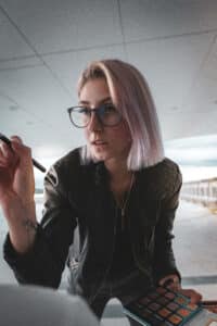 Eine Person mit schulterlangem Haar und Brille trägt sorgfältig Make-up mit einem Pinsel auf und hält eine Lidschattenpalette in der Hand. Sie trägt eine schwarze Lederjacke und bereitet sich in einem geräumigen, lichtdurchfluteten Raum auf eine Hochzeit vor.