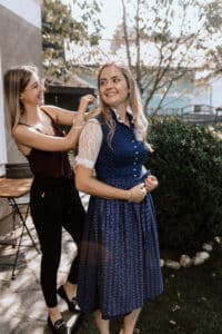 Zwei Frauen stehen draußen in einem Garten und sind in ihre Hochzeitsplanung vertieft. Eine rückt das lange, blau gemusterte Kleid der anderen zurecht. Sie lächeln beide, in der Nähe stehen ein Holzstuhl und ein Tisch, im Hintergrund sind grüne Büsche zu sehen. Es ist ein sonniger Tag, perfekt für die Planung einer Traumhochzeit.