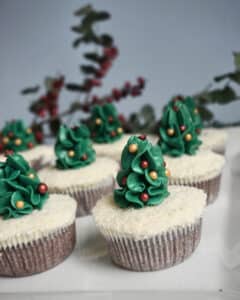 Cupcakes mit grünem Zuckerguss in Form von Weihnachtsbäumen, geschmückt mit essbaren roten und gelben Perlen, angeordnet auf einer weißen Oberfläche, die an eine Hochzeit erinnert. Im Hintergrund unterstreichen verschwommenes Grün und rote Beeren das festliche Thema und verleihen Ihrer Hochzeitsplanung einen Hauch Romantik.