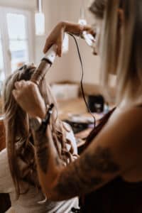 Eine Friseurin mit Tattoos auf dem Arm lockt das lange blonde Haar einer Frau mit einem Lockenstab. Die Kulisse ähnelt einem Salon und fängt die Aufregung der Hochzeitsplanung perfekt ein. Natürliches Licht strömt durch das Fenster und beleuchtet die Vorbereitungen für den Hochzeitstag.