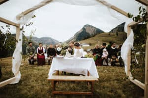 Ein Paar sitzt während einer Hochzeitszeremonie im Freien mit Bergen im Hintergrund an einem Tisch unter einem dekorativen Bogen. Gäste in traditioneller Kleidung sitzen auf Bänken. Die Umgebung hat eine rustikale, natürliche Atmosphäre, perfekt für diejenigen, die mitten in der Hochzeitsplanung stecken.