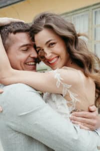 Ein fröhliches Paar umarmt sich während seiner Hochzeit, wobei die Frau breit lächelt. Sie trägt ein weißes Kleid mit Spitzendetails und hat welliges braunes Haar. Der Mann in einem hellgrauen Anzug hält sie fest, während sie einen glücklichen Moment vor einem Gebäude mit blassgrünen Verzierungen teilen.