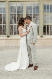Eine Braut in einem weißen Kleid und ein Bräutigam in einem hellgrauen Anzug stehen eng beieinander auf einem Kiesweg vor einem Gebäude mit großen Fenstern. Die Braut hält einen Blumenstrauß, während sie einander anschauen. Dies schafft eine romantische Hochzeitsatmosphäre, die wunderschön eingefangen wurde und exquisite Hochzeitsplanung widerspiegelt.