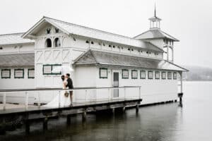 Braut und Bräutigam umarmen sich an einem regnerischen Tag unter einem Regenschirm auf einem Pier neben einem weißen, alten Bootshaus mit aufwendigen Verzierungen. Das ruhige Wasser spiegelt die heitere Atmosphäre ihrer Hochzeit wider und macht sie zu einem unvergesslichen Moment auf ihrer Hochzeitsreise.