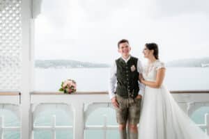 Ein Paar in traditioneller Kleidung steht auf einem überdachten Deck und verkörpert die Essenz einer Hochzeit. Mit Blick auf den ruhigen See und die Hügel trägt die Frau ein weißes Kleid, während der Mann eine Weste und Shorts trägt. Ein Blumenstrauß ruht auf dem Geländer in der Nähe, alle Elemente harmonieren mit der sorgfältigen Hochzeitsplanung.