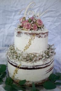 Eine rustikale, zweistöckige Naked Cake, perfekt für die Hochzeitsplanung, ist mit rosa Rosen und Schleierkraut geschmückt. Gekrönt wird sie von einem hölzernen „Love“-Schild und umgeben von grünen Eukalyptusblättern. Goldflocken betonen den strukturierten weißen Zuckerguss – ein Traum für alle, die heiraten möchten.