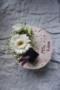 Ein runder Kuchen mit hellvioletter Glasur, perfekt für eine Hochzeit, gekrönt mit einer weißen Gerbera, weißen Rosen, kleinen weißen Blumen, silbernen Perlen und einer schwarzen Abschlusskappe als Dekoration. Die Worte „MSc. Lisa“ sind elegant auf den Kuchen geschrieben.