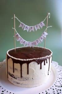 Ein glasierter Kuchen mit Schokoladenganache am Rand, gekrönt mit einem kleinen „Happy Birthday“-Banner. Der Kuchen ist ideal für eine Hochzeit, er ist mit einem perlenartigen Rand verziert und steht auf einem weißen Tortenständer mit Spitzenmuster vor einem grünen Hintergrund.