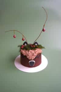 Eine dekorative Torte in Rentierform, perfekt für eine Winterhochzeit, mit Schokoladenglasur mit rosa Schnörkeln, geweihartigen Zweigen mit Ornamenten und Ohrdekorationen. Ideal, um Ihrer Hochzeitsplanung vor einem üppigen grünen Hintergrund eine skurrile Note zu verleihen.