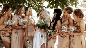 Eine Braut steht an ihrem Hochzeitstag unter einem Baum, umgeben von sechs Brautjungfern in passenden hellbraunen Kleidern. Sie lächeln alle und halten kleine Sträuße aus weißen Blumen in den Händen. Die Szene spielt im Freien an einem sonnigen Tag und fängt die Freude dieses wunderschön geplanten Anlasses ein.