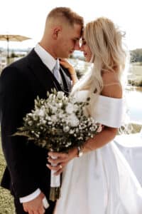 Braut und Bräutigam stehen in einem zärtlichen Moment ihrer Hochzeit eng beieinander und berühren ihre Stirnen in einer wunderschönen Umgebung im Freien. Die Braut hält einen Strauß weißer Blumen in der Hand und trägt ein schulterfreies Kleid. Der Bräutigam trägt einen schwarzen Anzug mit Krawatte. Beide lächeln und schaffen eine romantische Atmosphäre.