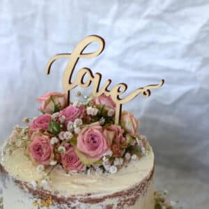 Eine rustikale Torte, perfekt für eine Hochzeit, verziert mit rosa Rosen und weißen Schleierkrautblüten. Ein hölzerner „Love“-Aufsatz schmiegt sich zwischen die Blüten. Die Torte hat eine strukturierte, halbnackte Glasuroberfläche mit einem dezenten Goldakzent an der Seite, der Ihrer Hochzeitsplanung Charme verleiht.