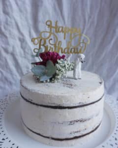 Ein halbnackter Kuchen mit weißem Zuckerguss ist mit „Happy Birthday“ in Gold, einer kleinen weißen Hundefigur und einem Strauß roter und weißer Blumen verziert, die an elegante Hochzeitsarrangements erinnern. Er wird wunderschön auf einem weißen Kuchenständer mit Spitzenrand präsentiert.