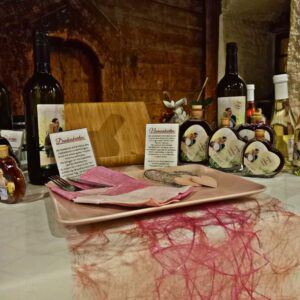 Eine Präsentation mit Weinflaschen, Marmeladengläsern und Informationskarten passend für eine romantische Hochzeit. Ein rosa Teller mit dekorativem Papier und ein Holzständer stehen vor einem rustikalen Holzhintergrund. Reichhaltige Texturen und Farben schaffen eine einladende, handwerkliche Atmosphäre, die perfekt für die Hochzeitsplanung ist.
