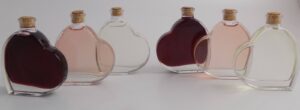 Herzförmige Glasflaschen mit Korken, gefüllt mit verschiedenfarbigen Flüssigkeiten – dunkelrot, hellrosa, klar und noch einmal dunkelrot – sind elegant auf einem weißen Hintergrund angeordnet. Perfekt, um Ihrer Hochzeit einen Hauch Romantik zu verleihen oder als charmanter Akzent in der Hochzeitsplanungsdekoration.