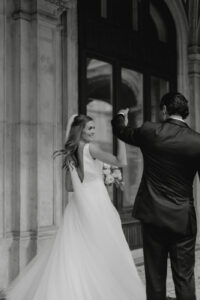 Braut und Bräutigam tanzen im Hochzeitskleid fröhlich vor einem großen Gebäude und feiern ihre Hochzeit. Das fließende weiße Kleid und der Schleier der Braut bilden einen schönen Kontrast zum dunklen Anzug des Bräutigams, der sie anmutig wirbelt. Der zeitlose Moment ist in Schwarzweiß festgehalten und verkörpert perfekte Hochzeitsplanung.