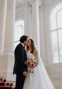 Eine Braut in weißem Kleid und Schleier küsst einen Bräutigam im schwarzen Anzug auf einer großen Treppe und verkörpert die Romantik des Heiratens. Sie hält einen Strauß rosa und weißer Blumen in den Händen, eingerahmt von großen Bogenfenstern und eleganten Säulen, die die Essenz exquisiter Hochzeitsplanung einfangen.