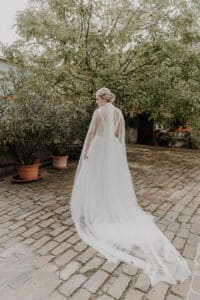 Eine Braut in einem langen, fließenden weißen Kleid und Schleier steht auf einem Kopfsteinpflasterweg, umgeben von Topfpflanzen und Bäumen, und verkörpert die Essenz einer idyllischen Hochzeit.