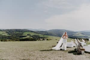 Eine Braut in einem weißen Kleid steht im Freien und hält einen Blumenstrauß in der Hand. Im Hintergrund sieht man sanfte grüne Hügel und einen teilweise bewölkten Himmel. In der Nähe stehen Reihen leerer weißer Stühle, perfekt für die Hochzeit arrangiert, und zeigen eine durchdachte Hochzeitsplanung.