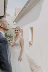 Eine Braut in einem eleganten weißen Kleid lächelt strahlend und hält einen Blumenstrauß in der Hand, was die Freude einer Hochzeit verkörpert. Sie steht neben einem weißen Gebäude mit einem verschwommenen Mann im Vordergrund. Die Außenkulisse ist hell und heiter und fängt die Essenz einer Hochzeit ein.