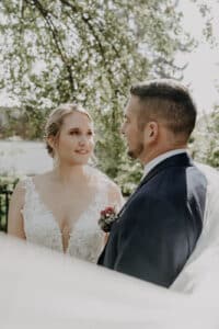 Braut und Bräutigam stehen im Freien und schauen sich unter einem sonnenbeschienenen Baum an. Sie verkörpern die Freude ihrer Hochzeit. Die Braut trägt ein weißes Spitzenkleid und der Bräutigam einen dunklen Anzug mit einer Blumenboutonniere. Der unscharfe Hintergrund unterstreicht den bezaubernden Moment ihrer Hochzeit.