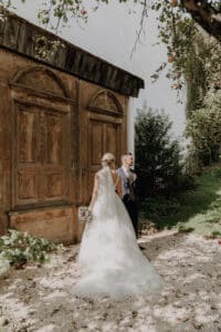 Braut und Bräutigam stehen draußen vor großen, rustikalen Holztüren. Während sie ihre Hochzeit feiern, trägt die Braut ein weißes Kleid und einen Schleier und hält einen Blumenstrauß, während der Bräutigam einen Anzug trägt. Umgeben von Grün fällt Sonnenlicht durch die Zweige darüber.