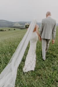 Braut und Bräutigam gehen Hand in Hand durch eine grüne Wiese und feiern ihre Hochzeit. Die Braut trägt ein weißes Spitzenkleid und einen langen Schleier, während der Bräutigam einen hellgrauen Anzug trägt. Sanfte Hügel bilden die Kulisse unter einem sanften, bewölkten Himmel – eine perfekte Szene für ihre Hochzeitsreise.
