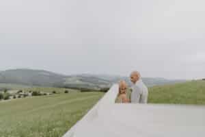 Braut und Bräutigam stehen zusammen auf einer Wiese mit sanften Hügeln im Hintergrund und feiern ihre Hochzeit. Der Schleier der Braut weht hinter ihnen her und erzeugt eine sanfte Unschärfe im Vordergrund. Der bewölkte Himmel verleiht diesem malerischen Moment der Hochzeit eine heitere Atmosphäre.
