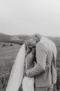 Ein Schwarzweißfoto zeigt ein Paar, das sich auf einem Feld umarmt und küsst. Die Frau in ihrem Hochzeitskleid mit langem Schleier und der Mann in seinem eleganten Anzug spiegeln die Essenz von Heiraten wider. Sanfte Hügel bilden eine atemberaubende Kulisse für diesen eleganten Moment.