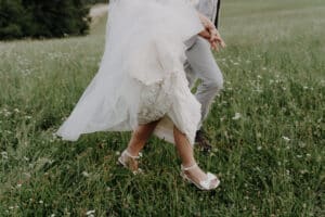 Ein Paar schlendert über eine Wiese und ist ganz in den Moment seiner Hochzeit vertieft. Das weiße Kleid der Braut fließt elegant, während sie in ihren weißen High Heels hervortritt, während der Bräutigam sie in hellen Hosen und dunklen Schuhen ergänzt. Wildblumen verleihen dieser zauberhaften Kulisse Charme, perfekt für jeden Hochzeitsplanungstraum.