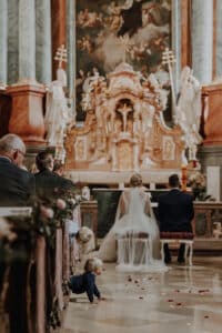 Eine Hochzeitszeremonie in einer großen Kirche. Braut und Bräutigam knien vor einem reich verzierten Altar, der mit Statuen und Kunstwerken geschmückt ist. Die Gäste sitzen und ein Kind krabbelt verspielt den Gang entlang, der mit Rosenblättern übersät ist.