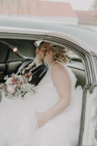 Ein frisch verheiratetes Paar küsst sich in einem Oldtimer und verkörpert damit die Romantik, von der jede Hochzeit träumt. Die Braut in ihrem weißen Kleid, einen Strauß rosafarbener und weißer Blumen umklammernd, sitzt neben dem Bräutigam in seinem dunklen Anzug und schafft so einen intimen Moment, der perfekt für ihre Hochzeitsreise ist.