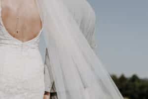 Eine Braut in einem rückenfreien, geblümten Spitzenbrautkleid steht neben einer Person in einem hellgrauen Anzug während ihrer Hochzeit. Ihr Schleier weht elegant im Wind. Der Himmel ist klar und im Hintergrund ist verschwommenes Grün zu sehen, das diesen wunderschönen Moment der Hochzeit einfängt.