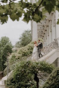 Braut und Bräutigam stehen gemeinsam auf einer kunstvoll verzierten, geschwungenen Treppe vor einem großen Gebäude. Üppiges Grün umgibt sie und schafft eine ruhige und romantische Atmosphäre, die perfekt für jede Hochzeitsplanung ist.