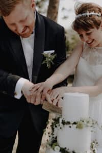 Ein Paar im Hochzeitskleid strahlt, als sie gemeinsam eine runde, weiße Hochzeitstorte anschneiden. Der Bräutigam trägt einen schwarzen Anzug und die Braut ein weißes Kleid, das mit einem Blumenkopfschmuck geschmückt ist. Die wunderschön gefertigte Torte ist mit üppigem Grün und zarten weißen Blumen dekoriert.