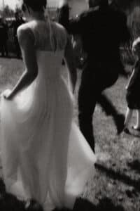 Schwarzweißfoto einer Braut in einem fließenden weißen Kleid, die über Gras läuft und die fröhliche Stimmung der Hochzeit einfängt. Neben ihr erscheint ein Mann im Anzug mitten im Sprung, während ein Kind fasziniert zusieht. Sonnenlicht wirft verspielte Schatten auf die Szene.