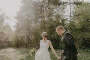 Braut und Bräutigam gehen Händchen haltend durch einen sonnenbeschienenen Garten und genießen die Freude ihrer Hochzeit. Die Braut trägt ein weißes Kleid und hält einen kleinen Blumenstrauß, während der Bräutigam einen schwarzen Anzug trägt. Bäume und Grün umgeben sie, in sanftes Sonnenlicht getaucht, und fangen die Essenz ihrer Hochzeit ein.