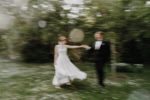 In einer verträumten, ätherischen Atmosphäre fängt ein verschwommenes Bild ein Paar ein, das während seiner Hochzeit in einem Garten tanzt. Die Frau gleitet in einem fließenden weißen Kleid dahin, während der Mann sie in einem eleganten schwarzen Anzug ergänzt, umgeben von üppigem Grün und sanftem Licht.