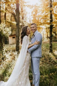 Braut und Bräutigam feiern ihre Hochzeit, indem sie sich in einem sonnenbeschienenen Herbstwald umarmen und lächeln. Die Braut trägt ein weißes Spitzenkleid und der Bräutigam einen hellblauen Anzug. Goldene Blätter umgeben sie und schaffen eine warme und romantische Atmosphäre, perfekt zum Heiraten.