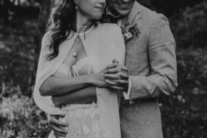 Ein Schwarzweißfoto zeigt ein umarmendes Paar an seinem Hochzeitstag. Die Frau trägt ein elegantes Spitzenkleid mit Schal und der Mann einen hellen Anzug mit einer Ansteckblume. Sie halten Händchen und lächeln, umgeben von einem unscharfen natürlichen Hintergrund.