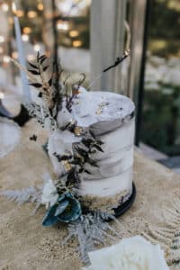 Eine rustikale, zweistöckige Hochzeitstorte mit strukturiertem weißen und grauen Zuckerguss ist ein schönes Element jeder Hochzeit. Geschmückt mit getrockneten Blumen und Blättern in neutralen Tönen sitzt sie auf einer goldbeigen Oberfläche und verleiht Ihrer Hochzeitsplanung einen charmanten Hintergrund aus Lichtern und Grün.