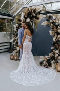 Ein Paar umarmt sich in einem eleganten Ambiente und fängt damit die Essenz von „Heiraten“ perfekt ein. Die Frau trägt ein detailreiches Spitzenkleid mit langer Schleppe, während der Mann einen hellblauen Anzug trägt. Sie stehen inmitten dekorativer Blumenbögen mit rustikalen Elementen in einem mit Glaswänden versehenen Veranstaltungsort.