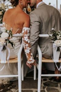 Braut und Bräutigam sitzen eng beieinander auf weißen Stühlen, geschmückt mit Rosen und Bändern, ein Beweis für exquisite Hochzeitsplanung. Händchenhaltend drehen die Braut in Blumenspitze und der Bräutigam im hellgrauen Anzug ihre Köpfe zueinander und genießen den intimen Moment des Heiratens.