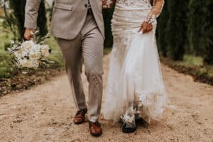 Braut und Bräutigam schlendern einen Sandweg entlang und verkörpern so ihren Hochzeitsmoment. Der Bräutigam in einem hellgrauen Anzug hält einen Strauß weißer Blumen, während die Braut anmutig den Saum ihres weißen Spitzenkleides hebt. Bäume säumen ihren Weg und rahmen diese wunderschöne Feier ein.