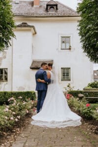 Ein Paar in Brautkleid und Anzug umarmt sich auf einem von Blumen umgebenen Weg, im Hintergrund ein großes weißes Gebäude. Grün umrahmt die Szene und schafft eine romantische Hochzeitskulisse, die wie aus einem Märchen erscheint.