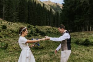 Auf einer üppigen, grünen Waldlichtung mit majestätischen Bergen im Hintergrund stehen Braut und Bräutigam bereit zum Heiraten. Die Braut hält einen Strauß blauer Blumen in der Hand, während sie sich lächelnd einander entgegenstrecken. Beide tragen leichte und legere Hochzeitskleidung.