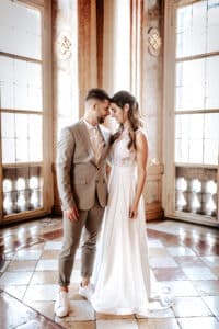 Braut und Bräutigam stehen eng beieinander in einem eleganten Raum mit großen Fenstern und kunstvollem Dekor und verkörpern die Essenz einer Traumhochzeit. Der Bräutigam trägt einen hellgrauen Anzug mit weißem Hemd und Turnschuhen, während das lange weiße Kleid der Braut blendet. Sie blicken sich liebevoll an.